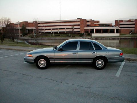 1995 Ford Crown Victoria - A silver 1995 Ford Crown Victoria sedan is parked in a lot with a building and water in the background.