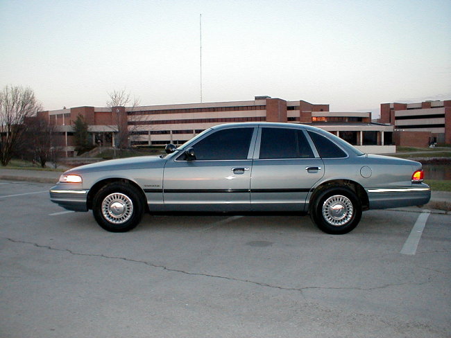 Light blue Ford Crown Victoria sedan, driver-side profile, parked in lot by brick building