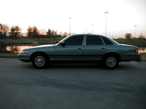 1995 Ford Crown Victoria - A 1995 Ford Crown Victoria sedan is seen from the side, parked near a body of water with trees in the background.