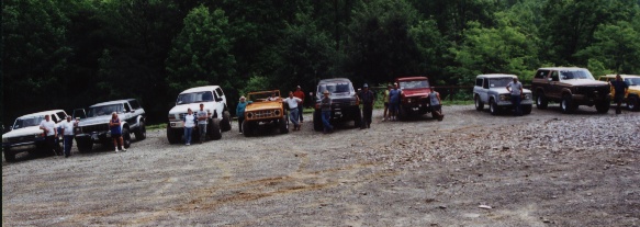 Row of Ford Broncos and trucks parked in a gravel lot with people standing nearby, trees behind