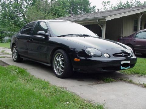 1999 Ford Taurus - A black 1999 Ford Taurus is parked in front of a house, with another vehicle visible in the background.