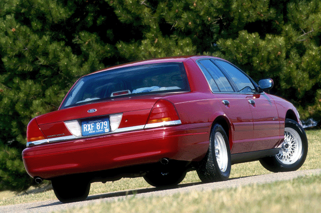 Dark red full-size sedan rear three-quarter view with chrome wheels on grass-lined road