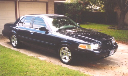Dark blue Ford Crown Victoria sedan, front three-quarter view, parked on driveway by brick house