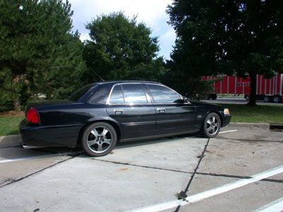 2001 Ford Crown Victoria - Black Ford Crown Victoria full-size sedan seen from the rear driver's side, fitted with polished multi-spoke aftermarket wheels, parked in…