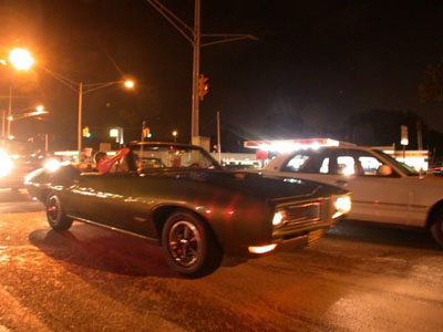 2001 Ford Crown Victoria - Night cruise shot of a dark first-generation Pontiac Firebird convertible with the top down and red interior, rolling on a wet-looking stre…