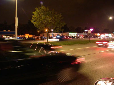 2001 Ford Crown Victoria - Blurred night street scene of a dark car passing in the foreground with motion-streaked taillights, shot along a multi-lane road lined with…