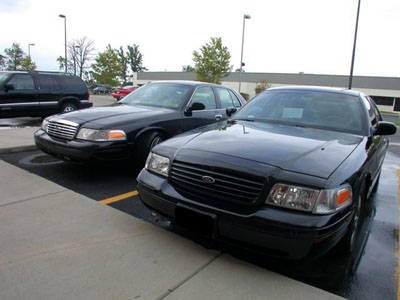 2001 Ford Crown Victoria - Two black Ford full-size sedans parked side by side in a wet parking lot, shot from the 3/4 front with blacked-out grilles, no front plates…