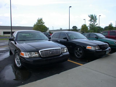 2001 Ford Crown Victoria - Two black Ford Crown Victoria sedans parked side by side in a wet lot, the near car on polished five-spoke wheels with a chrome grille and…