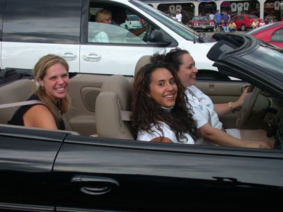 2001 Ford Crown Victoria - Three smiling women sit in a tan-interior black convertible at an outdoor car event, with a white SUV and other parked cars behind them.
