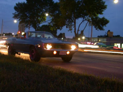 2001 Ford Crown Victoria - A dark first-generation Chevrolet Camaro convertible with the top down cruises a divided roadway at dusk, headlights and parking lamps lit…