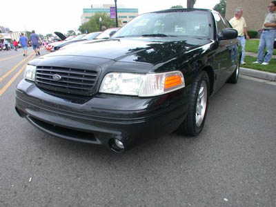 2001 Ford Crown Victoria - Black 1998+ Ford Crown Victoria at a car show, seen from a low front three-quarter angle. It has a blacked-out mesh grille with Ford oval,…