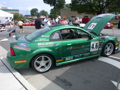 2001 Ford Crown Victoria - Green Ford Mustang race coupe at an outdoor car show with its hood open, shot from the 3/4 rear driver side on silver five-spoke wheels wit…