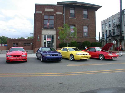 2001 Ford Crown Victoria - Four Ford Mustangs: red, dark blue, yellow and a red convertible with its hood up, lined up nose-out along a street in front of a two-story…