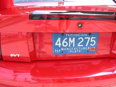 2001 Ford Crown Victoria - Close-up of a red car's decklid showing a blue Michigan "MANUFACTURER PLATE" reading 46M 275 with a FEB '04 tab, mounted below a body-color…