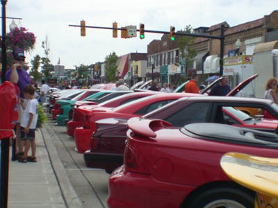 2001 Ford Crown Victoria - A long line of Mustangs and other muscle cars parked hood-up along a small-town main street cruise-in, viewed low from behind the rear spoi…