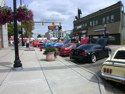2001 Ford Crown Victoria - Downtown street car show with vehicles parked at an angle along a sidewalk lined with hanging flower baskets and a brick storefront block.…
