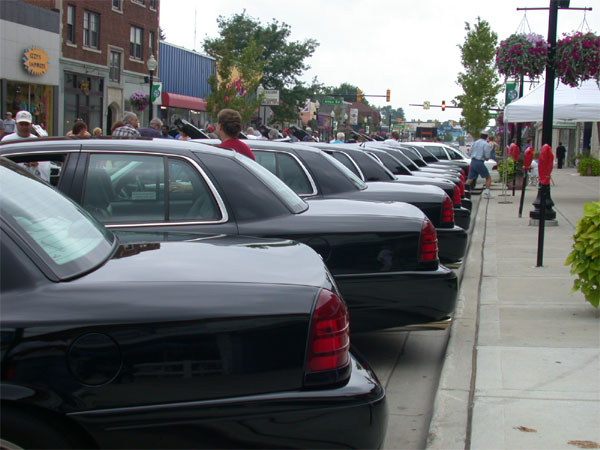 Row of black full-size sedans parked along a small-town street at a car show