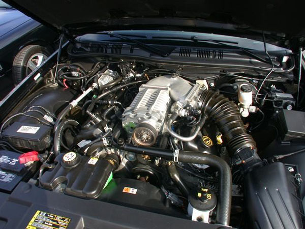 Engine bay with supercharged V8, black intake tubing and wiring under an open hood.