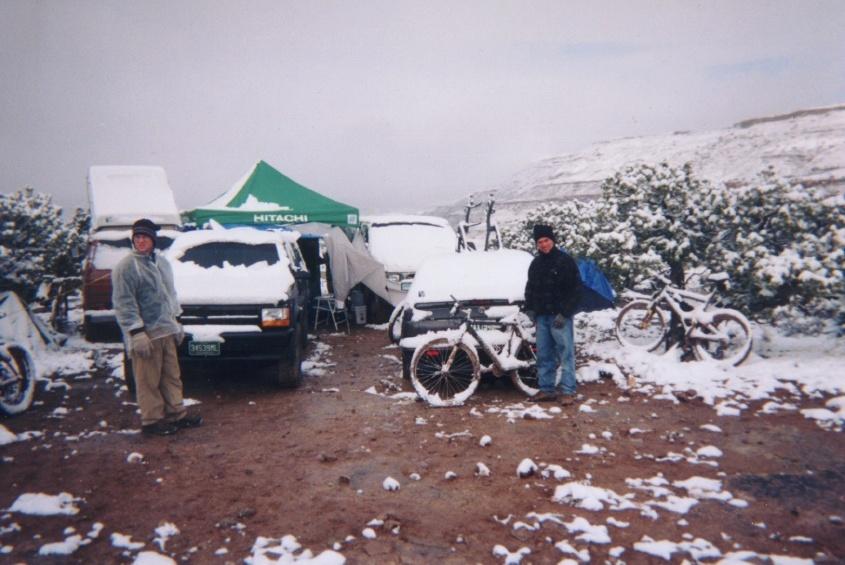 Snow-covered dark Ford SUV at a muddy campsite with green Hitachi tent, two people, and snowy mountain bikes