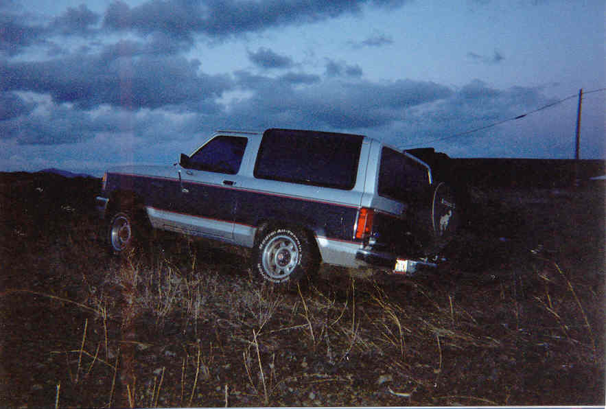Silver and blue two-door SUV parked in dry brush at dusk, rear three-quarter view