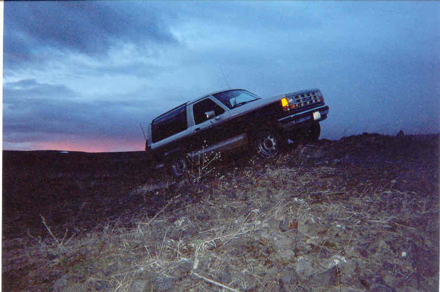Two-door SUV on rocky brush terrain at dusk, front 3/4 view with lights on