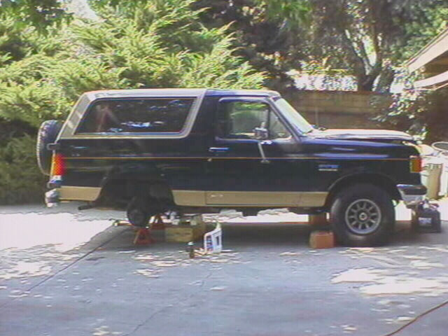Black and tan Ford Bronco SUV on jack stands in a driveway with rear wheel removed