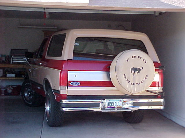 Red and tan Ford Bronco parked in a garage, rear view with Bronco tire cover