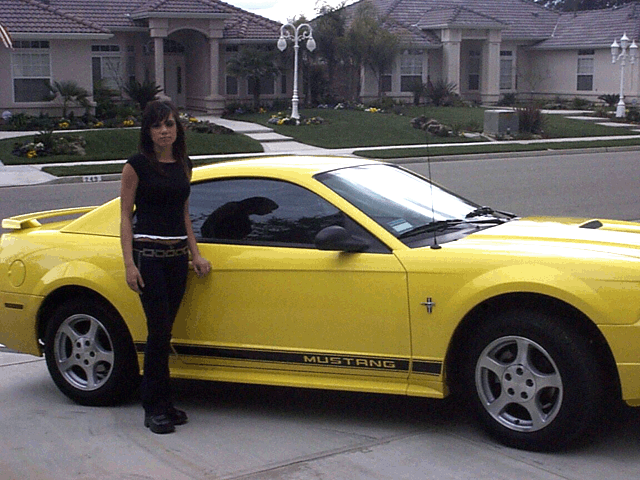 Yellow Ford Mustang coupe with black Mustang side stripe, woman standing beside it in a suburban driveway