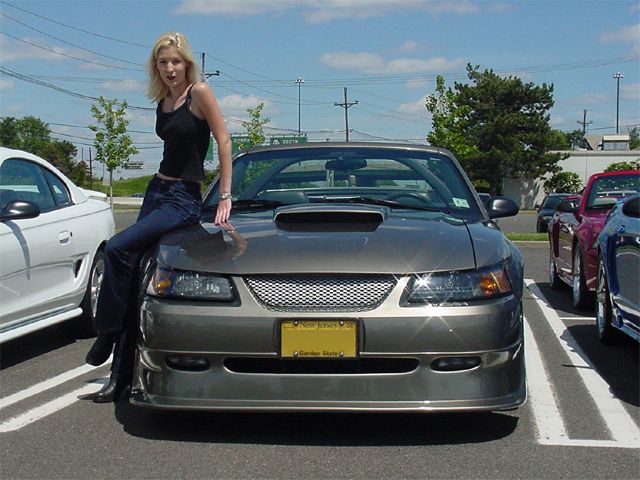 Woman sitting on hood of gray Ford Mustang convertible with hood scoop in parking lot
