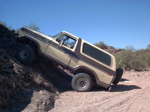 1979 Ford Bronco - A tan and brown two-tone 1979 Ford Bronco full-size SUV climbing a rocky embankment at an off-road trail, shown from a 3/4 front angle with…