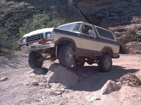 1979 Ford Bronco - A two-tone tan and brown 1979-era Ford Bronco full-size SUV climbing over a large rock on a rugged desert trail, shown from the front-left…