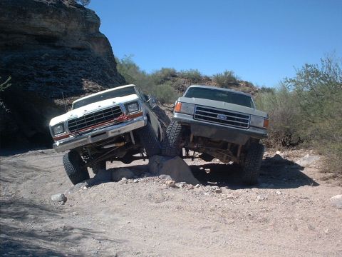 1979 Ford Bronco - Two Ford trucks climbing over rocks on a rocky off-road trail, both photographed from the front. The left vehicle is a white older-generati…