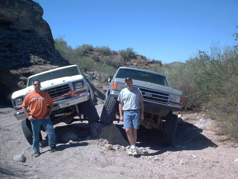 1979 Ford Bronco - Two lifted full-size Ford 4x4s parked nose-out on rocks in a desert wash with two men standing in front. The left truck is white with a red…