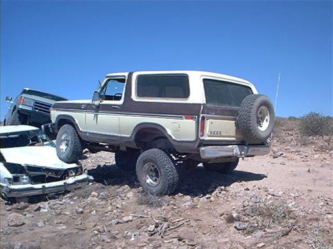 1979 Ford Bronco - A two-tone tan and brown 1979 Ford Bronco full-size SUV shown from the rear three-quarter, lifted on chunky off-road tires with a rear-moun…