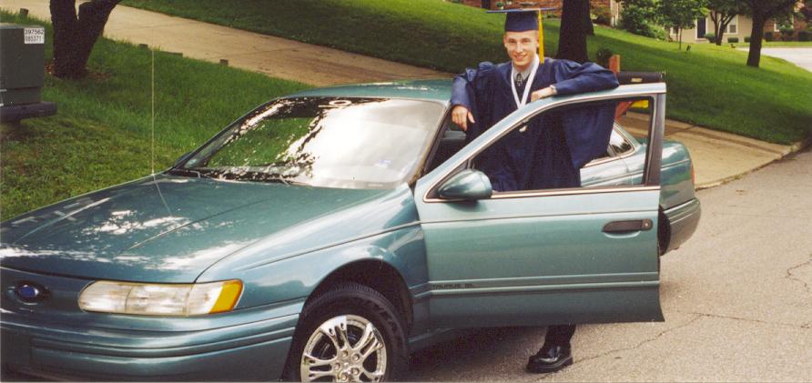 Teal Ford Taurus sedan with door open and graduate in blue cap and gown standing beside it
