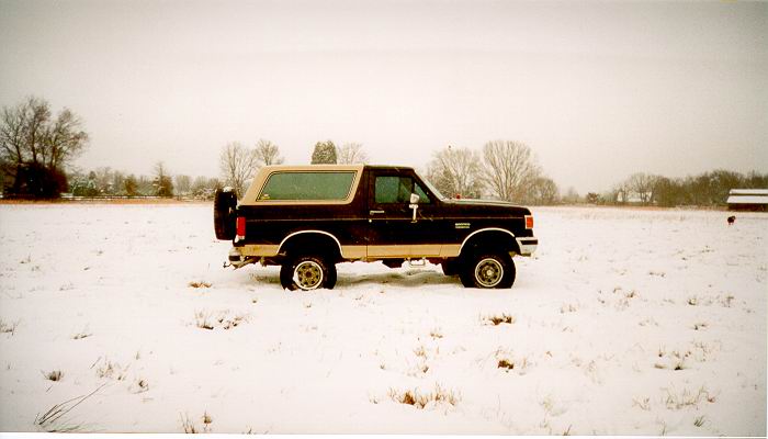 Black and tan Ford Bronco parked in a snowy field, side view with rear spare tire