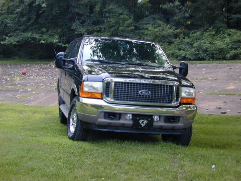 2001 Ford Excursion - This large SUV is parked on a grassy area with trees and a dirt path behind it.