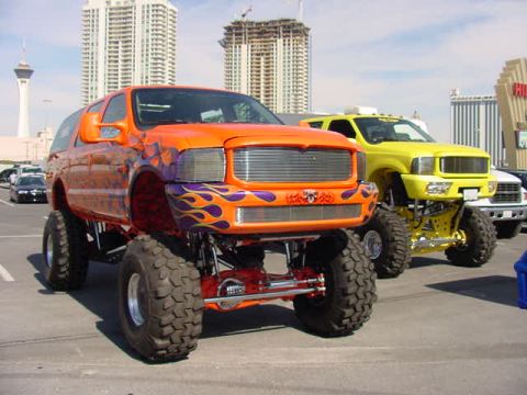 2001 Ford Excursion - Two lifted monster trucks, one orange with flames and one yellow, are parked on a street with buildings in the background.