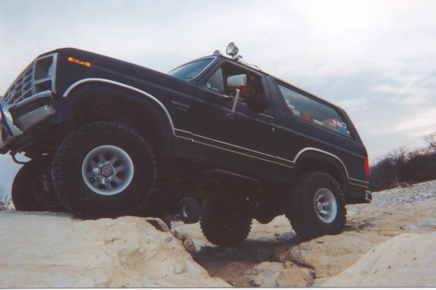 Lifted dark Ford Bronco on big tires crawling over rocks, low front-quarter view with driver visible