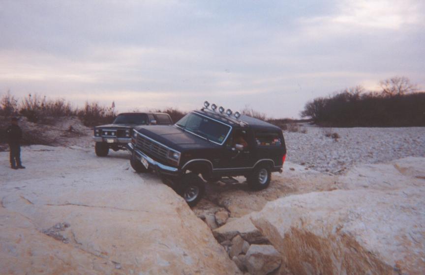 Lifted dark Ford Bronco with roof lights descending rocks off-road, second truck behind