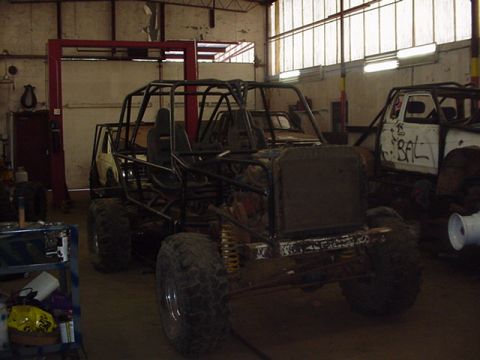 1979 Ford Bronco - A heavily modified off-road vehicle with a prominent roll cage is parked inside a workshop, with another vehicle visible in the background.