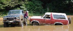 1979 Ford Bronco - Two off-road vehicles are stuck in murky water, with one person attempting to pull the red Bronco out with a blue truck.