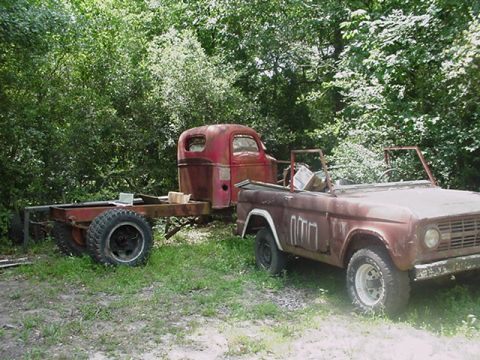 1979 Ford Bronco - Two old vehicles, a Ford Bronco and a truck chassis, sit in a grassy, wooded area.