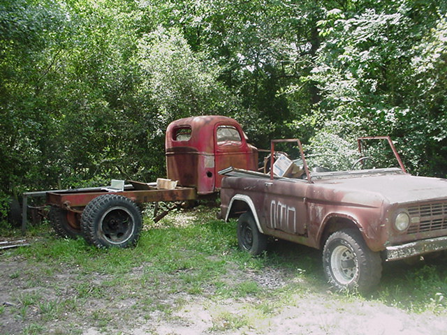 Rusty 1979 Ford Bronco next to an old red truck chassis with large tires