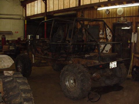 1979 Ford Bronco - Stripped 4x4 chassis under construction in a wood-walled shop, reduced to a bare rusty tub wrapped in a full tube cage with two bucket seat…
