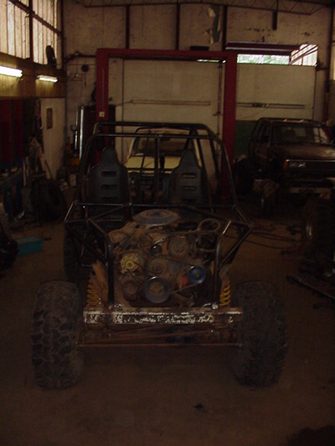 1979 Ford Bronco - Front view of a stripped tube-frame off-road buggy under construction inside a dim shop, with a full roll cage, two black plastic bucket se…