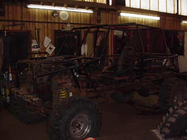 Stripped tube-cage trail rig on mud tires inside a wood-walled barn shop