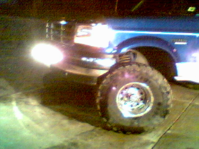 Lifted blue Ford SUV at night, headlights on, large mud tire with chrome wheel visible