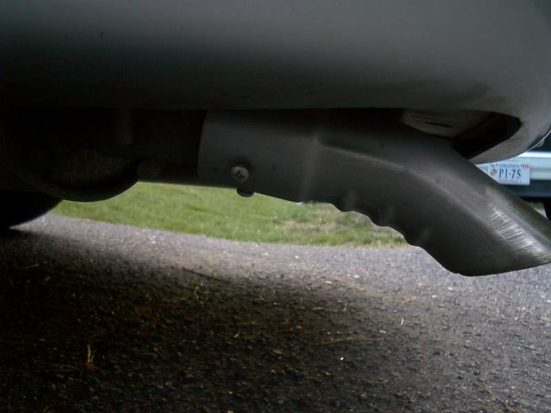 Close-up of a downturned chrome exhaust tip under a car's rear bumper, Virginia-plated car in background