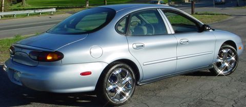 1998 Ford Taurus - This silver sedan is shown from the rear quarter angle, parked on a paved surface with a road and trees in the background.